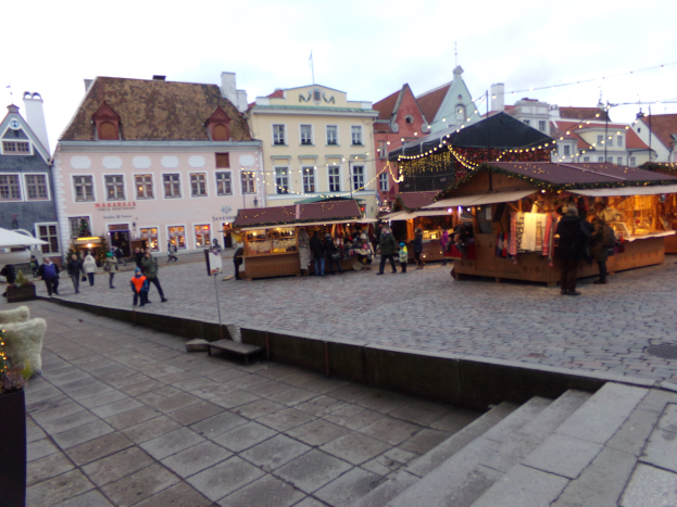 Ein geschäftiger Weihnachtsmarkt in Tallinn, Estland mit Menschen um geschmückte Stände, festliche Lichter, bewölkten Himmel, Stufen nach oben und Topfpflanzen im Bereich.