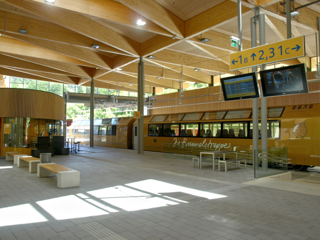 Ein belebter Bahnhof mit einem Zug auf den Gleisen, umgeben von Bänken, Tischen, Pfosten, Schildern, Laternen und Bäumen und voller Menschen, die auf den Zug warten.