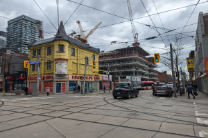 Ein gelbes Gebäude im Bau auf einer Stadtstraße mit Autos, Fußgängern, Ampeln, Strommasten, elektrischen Leitungen, Gebäuden, Schildern, Bäumen, Kränen und einem bewölkten Himmel.