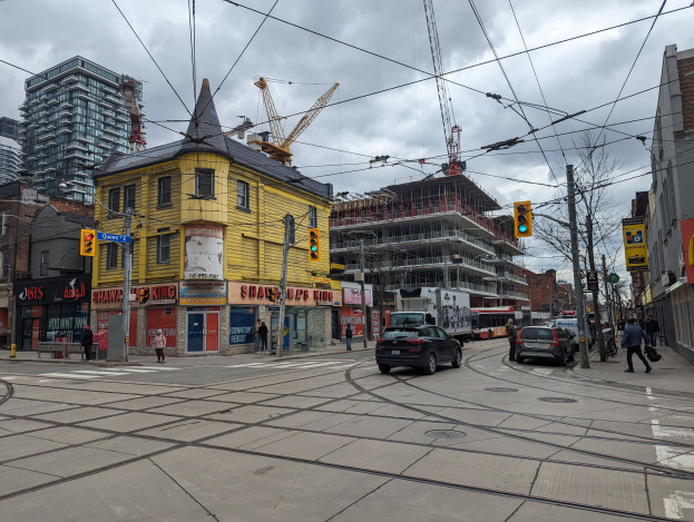 Ein gelbes Gebäude im Bau auf einer Stadtstraße mit Autos, Fußgängern, Ampeln, Strommasten, elektrischen Leitungen, Gebäuden, Schildern, Bäumen, Kränen und einem bewölkten Himmel.