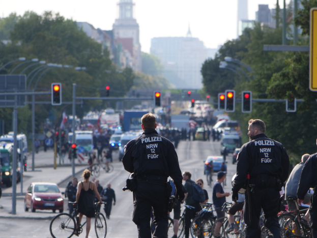 Polizeibeamte fahren mit Fahrrädern eine von Bäumen gesäumte Straße mit Gebäuden und einem klaren blauen Himmel im Hintergrund.
