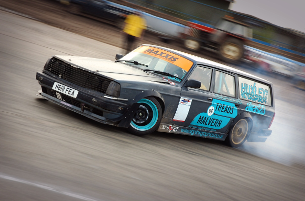 Ein Auto rast auf einer Rennstrecke, wobei Rauch ausstößt, umgeben von anderen Fahrzeugen und Zuschauern.