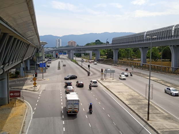 Eine stark befahrene Autobahn mit Schwerlastverkehr, neben einer Brücke, mit Straßenlaternen, Ampeln, Verkehrszeichen, Bäumen, Gebäuden, Hügeln und einem bewölkten Himmel im Hintergrund.