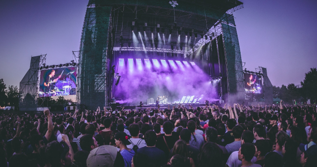 Eine große Menschenmenge auf einem Musikfestival schaut auf einer hell beleuchteten Bühne mit Instrumenten und Bildschirmen, die vor einem Gebäude, Bäumen und einem klaren blauen Himmel aufgebaut sind.