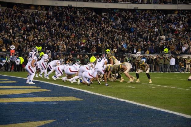 Ein Fußballspiel auf einem Stadionfeld mit Spielern in Sportbekleidung und Helmen auf dem Boden liegend, umgeben von Zuschauern, Geländern und Beschriftungen.