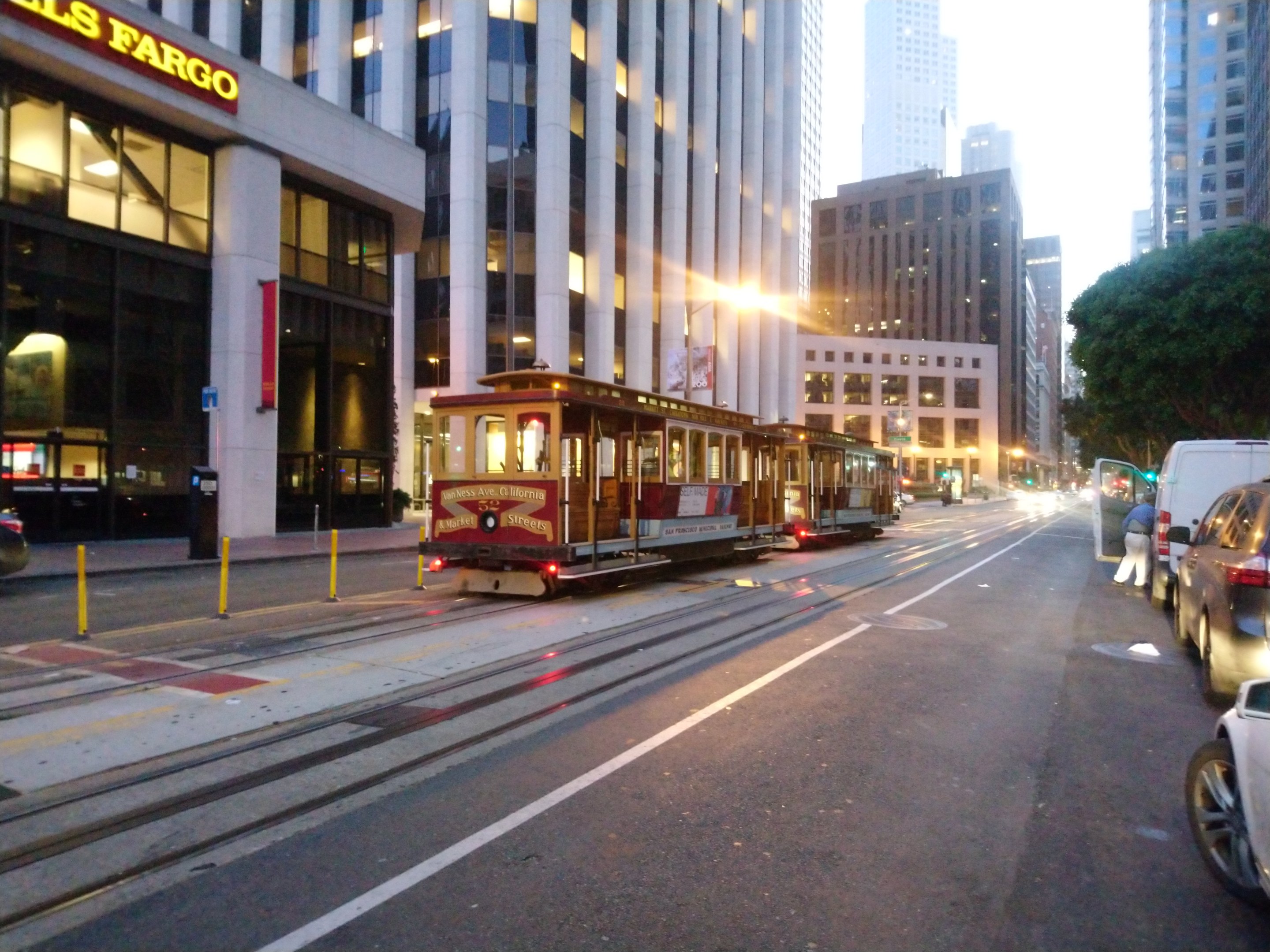 Eine Seilbahn fährt an einer Stadtstraße in San Francisco entlang, mit Fahrzeugen, einem Fußgänger und einem geparkten Van auf der Straße.