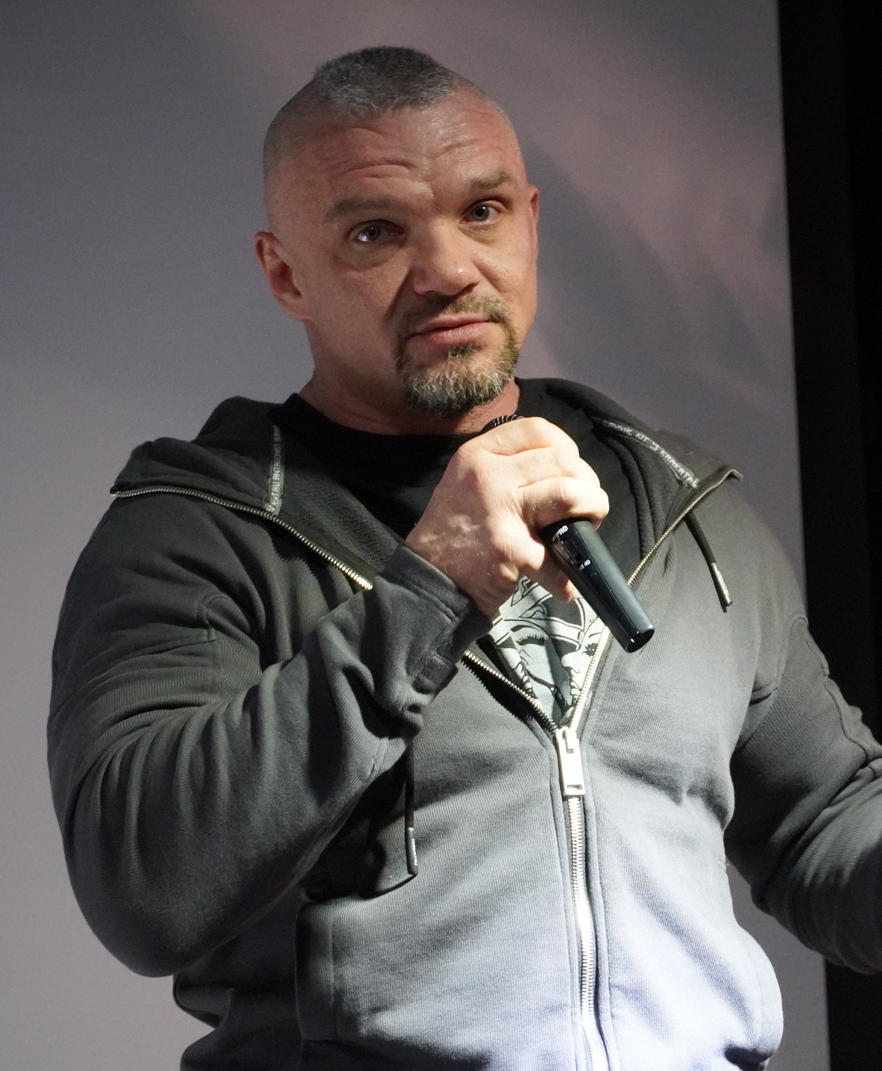 Ein Mann in einer schwarzen Kapuzenjacke und einem schwarzen Anzug mit einem weißen Hemd hält ein Mikrofon und steht selbstbewusst vor einer Wand, wobei er wie ein Komiker mit zurückgekämmtem Haar aussieht.