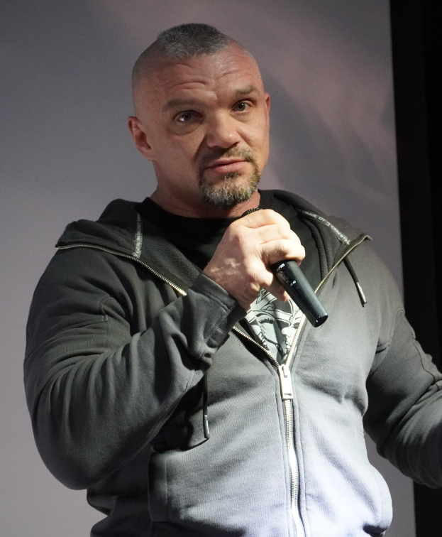 Ein Mann in einer schwarzen Kapuzenjacke und einem schwarzen Anzug mit einem weißen Hemd hält ein Mikrofon und steht selbstbewusst vor einer Wand, wobei er wie ein Komiker mit zurückgekämmtem Haar aussieht.