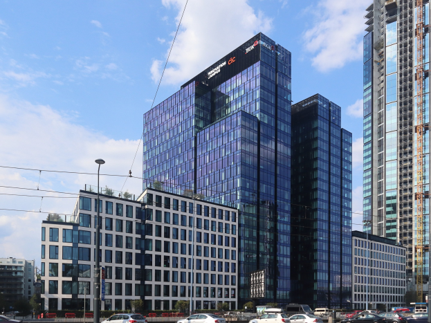Zentrale der Deutschen Bank in Frankfurt, Deutschland, ein großes Gebäude mit vielen Fenstern, umgeben von städtischen Elementen einschließlich Straßenlaternen, Fahrzeugen, Bäumen und einem bewölkten Himmel.