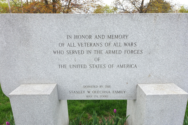 Denkmalstein mit einer Widmung an US-Streitkräfte-Veteranen, umgeben von Pflanzen, Blumen, Bäumen und einer bewölkten Himmel.