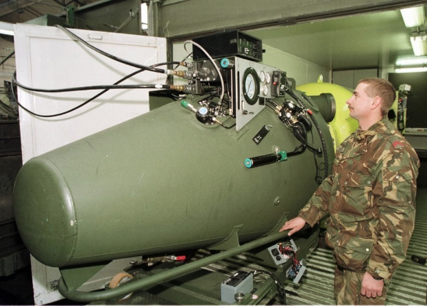Ein Mann in militärischer Uniform steht neben einer großen grünen Bombe in einer unordentlichen industriellen Umgebung mit Maschinen, Drähten und beleuchteten Wandlampen, mit Text unten.