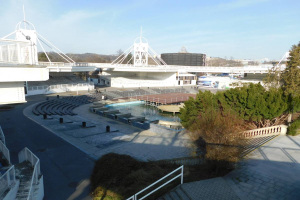 Blick auf das Olympische Stadion von einem hohen Punkt aus, mit verschiedenen Objekten und Grün im Vordergrund und einem bewölkten Himmel im Hintergrund.