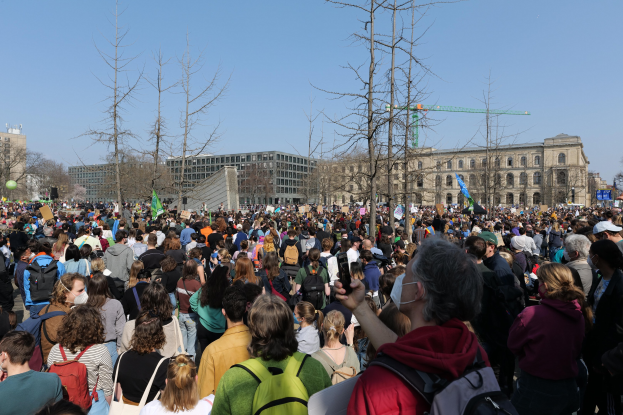 Große Menge von Menschen mit Schildern und Taschen vor einem Gebäude mit Bäumen und einem klaren Himmel, die an einer Klimawandel-Demonstration teilnehmen.