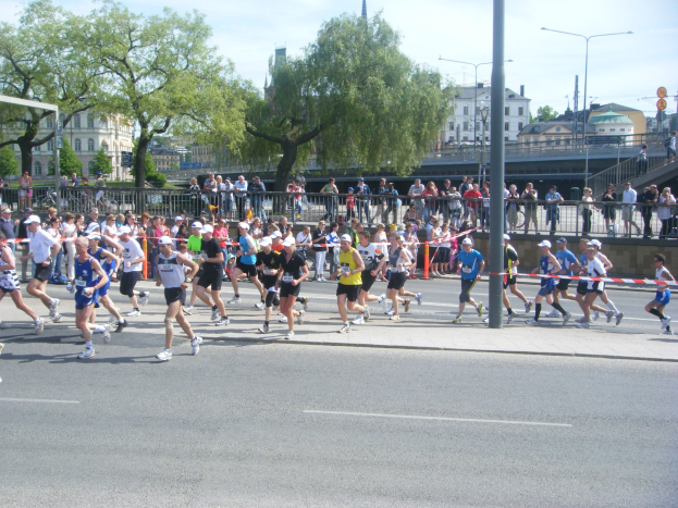 Gruppe von Läufern bei einem Marathon auf einer Straße mit Zielpfosten und Ribbon-Finish, umgeben von Metallzaun und Absperrungen, Zuschauern auf dem Gehweg, Pfosten, Schilder, Brücke, Gebäuden, Bäumen und bewölktem Himmel.
