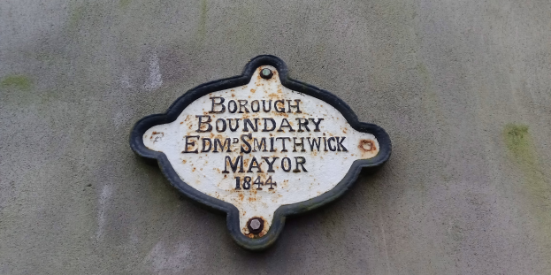 Metallplatte an einer Gebäudeseite mit schwarzem Hintergrund und weißer fetter Schrift, die "Gemeindegrenze Edm Smithwick Bürgermeister" liest.