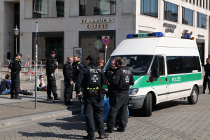 Eine Gruppe von Polizeibeamten steht vor einem Starbucks-Café, rechts daneben ein Van, links Menschen und im Hintergrund ein Gebäude mit Fenstern, einer Schautafel, einem Laternenmast und einem Zaun.