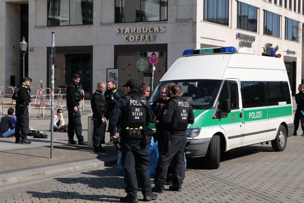 Eine Gruppe von Polizeibeamten steht vor einem Starbucks-Café, rechts daneben ein Van, links Menschen und im Hintergrund ein Gebäude mit Fenstern, einer Schautafel, einem Laternenmast und einem Zaun.