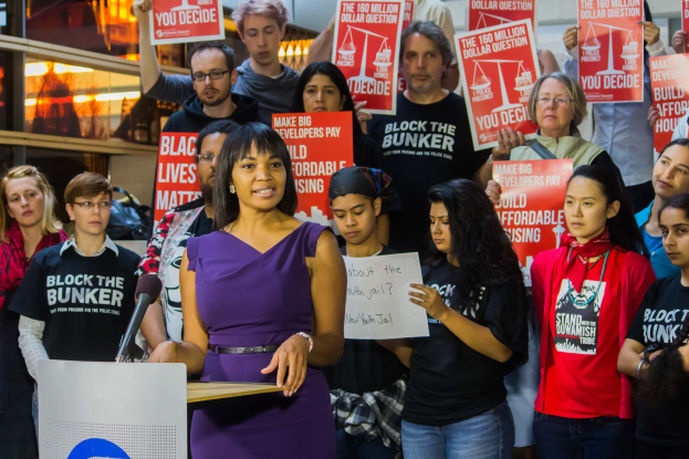 Eine Black Lives Matter-Aktivistin spricht an einem Rednerpult mit Mikrofon zu einer Menge mit Schildern, während im Hintergrund Lichter und Gegenstände zu sehen sind.