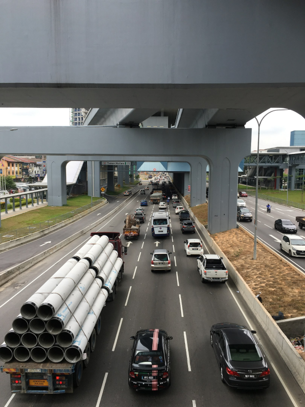 Eine vielbefahrene Autobahn mit zahlreichen Fahrzeugen, einer Brücke darüber, Straßenlaternen, Gras, Gebäuden, Bäumen und einem Himmel als Hintergrund.