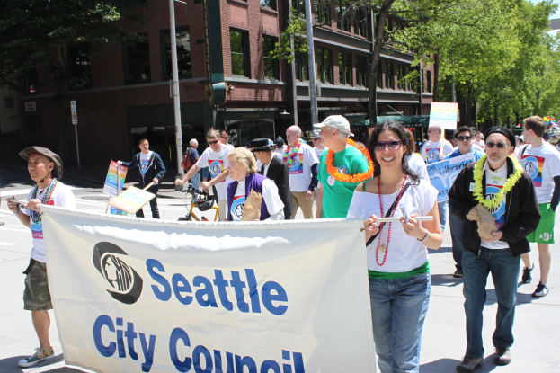 Eine Gruppe von Menschen marschiert mit einem 'Seattle City Council'-Schild die Straße entlang, einige tragen Mützen und Kränze, andere halten Schilder, mit Bäumen, Gebäuden, Laternenpfählen und einem klaren blauen Himmel im Hintergrund.