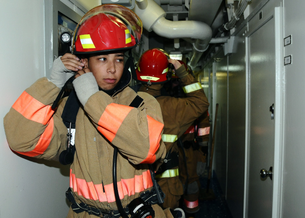 Feuerwehrleute in Uniform stehen in einem Raum mit einer Tür auf der rechten Seite und einer Wand auf der linken Seite während einer Übung, mit Rohren im Hintergrund.