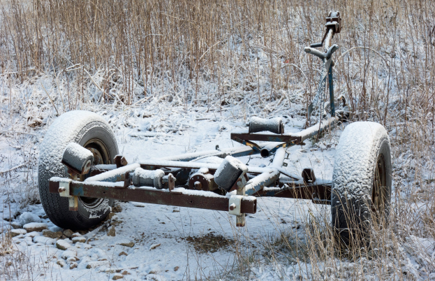 Alter rostiger Anhänger voller Schnee auf einem verschneiten Feld mit Gras und Steinen.