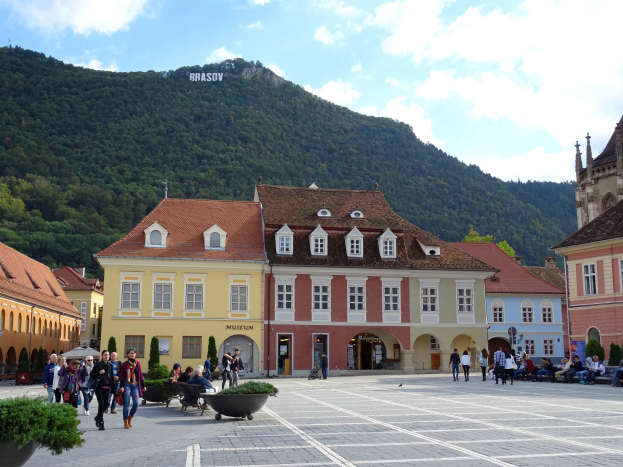 Menschen gehen auf einem Stadtplatz umgeben von Gebäuden mit Fenstern, Laternenmasten und Topfpflanzen, mit einem Hügel und Bäumen im Hintergrund unter einem bewölkten Himmel.