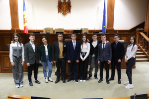 Eine Gruppe von Menschen, Gewinner des European Court of Justice 2018-2019, die in einem Gerichtssaal vor Monitoren, Mikrofonen und anderen Gegenständen auf Tischen in der Vordergrund und einer hölzernen Plattform mit Stühlen, Flaggen und einem Gegenstand an der Wand im Hintergrund lächeln und posen.