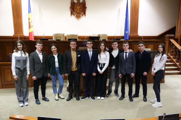 Eine Gruppe von Menschen, Gewinner des European Court of Justice 2018-2019, die in einem Gerichtssaal vor Monitoren, Mikrofonen und anderen Gegenständen auf Tischen in der Vordergrund und einer hölzernen Plattform mit Stühlen, Flaggen und einem Gegenstand an der Wand im Hintergrund lächeln und posen.