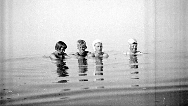 Schwarzes und weißes Foto von vier Kindern, die in einem Pool schwimmen, jedes trägt eine Schwimmkappe.