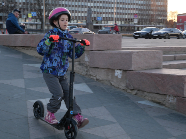 Ein junger Junge mit Helm und Handschuhen fährt auf einem Gehweg mit Stufen, Fahrzeugen, Menschen, Bäumen, Pfosten, Brettern, Gebäuden und einem klaren blauen Himmel im Hintergrund.