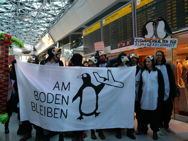Gruppe von Menschen in Pinguinkostümen, die ein Banner vor einer Wand mit Pinguin-Displays, Schaufensterpuppen und anderen Gegenständen halten.